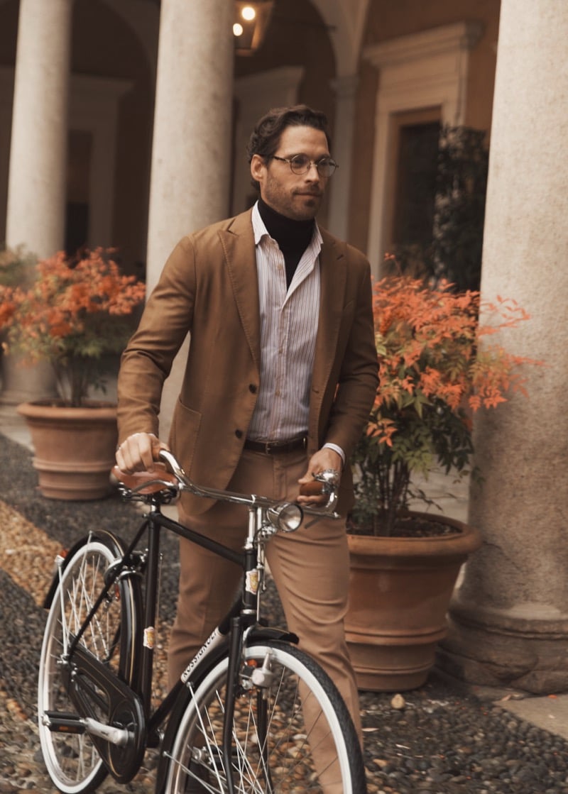 Simone Bredariol with a bicycle among autumn foliage in a camel suit over a dark turtleneck and striped shirt, for Boglioli's fall-winter 2026 campaign
