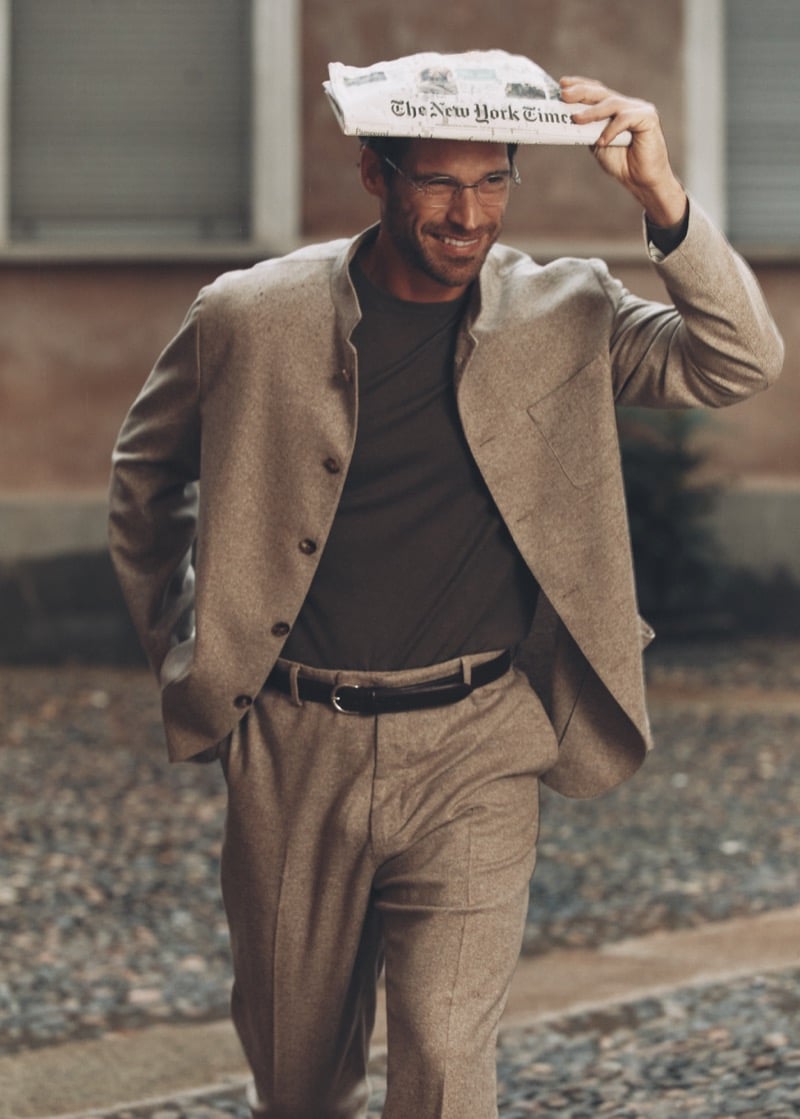 Simone Bredariol holding a newspaper over his head in a camel collarless suit and brown T-shirt on a Milan street, for Boglioli's fall-winter 2026 campaign