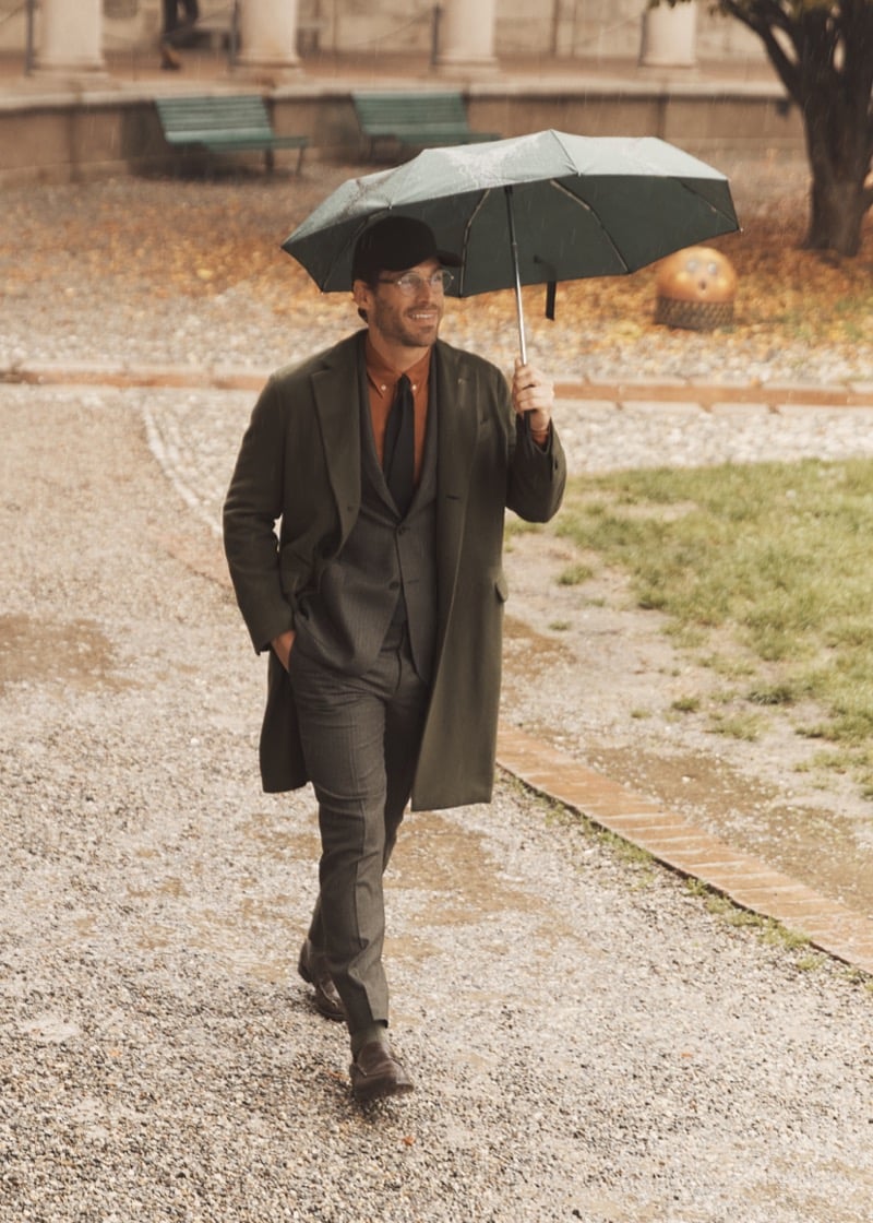 Simone Bredariol walking in the rain with a green umbrella in a green overcoat over a gray three-piece suit, for Boglioli's fall-winter 2026 campaign