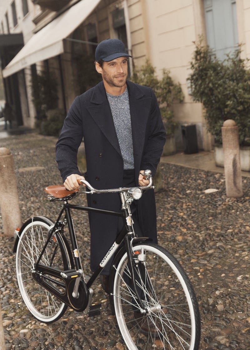 Simone Bredariol with a vintage bicycle in a dark peacoat over a marled gray sweater and a cap, for Boglioli's fall-winter 2026 campaign