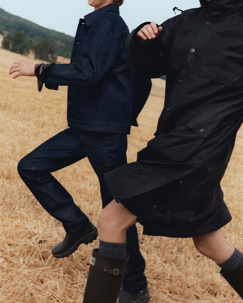 Joseph Uyttenhove in dark denim walking through a field beside another model in the Barbour x A.P.C. campaign.