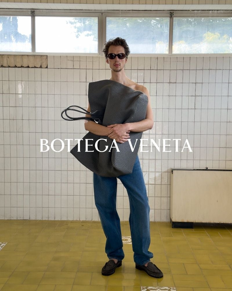 Leon Dame in sunglasses and jeans holding an oversized dark woven Bottega Veneta bag against his bare torso.