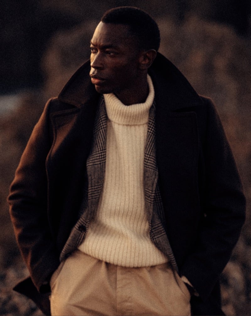 Akin Jaiye wears a peacoat over a checked jacket and ivory turtleneck with pleated chinos.