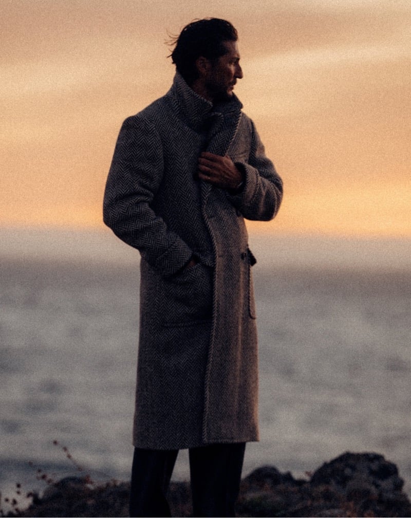 Ryan Porter wears a long herringbone coat beside the sea.