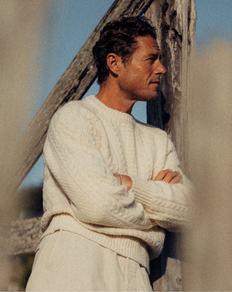 Jon Paul Phillips wears an ivory cable-knit sweater with light trousers beside a wooden post.