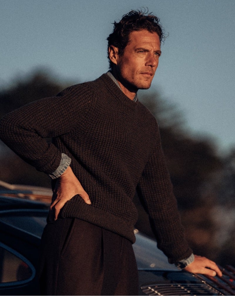 Jon Paul Phillips wears a thick brown sweater with pleated trousers beside a vintage car.