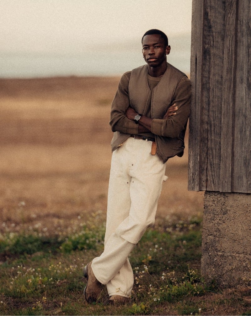 Akin Jaiye wears a knit polo and light trousers beside a wooden wall.