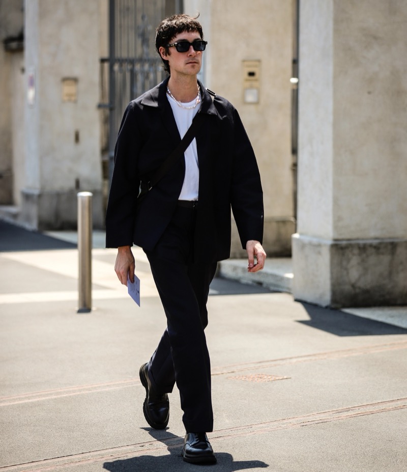 Man in a black jacket, white tee, black trousers and black leather shoes walking down a street in sunglasses