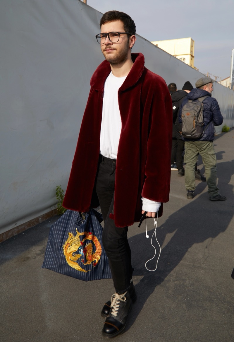 Man in a burgundy fur coat, white tee, black jeans and black boots carrying a striped bag with an embroidered dragon