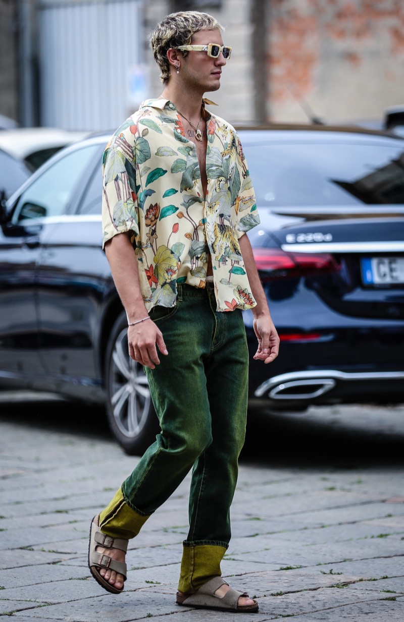 Man in a cream floral print shirt, green jeans with yellow cuffs and tan sandals on a cobblestone street