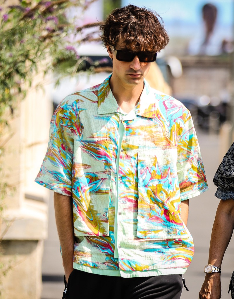 Man in a short-sleeve shirt with a bold multicolor print, black sunglasses and black shorts