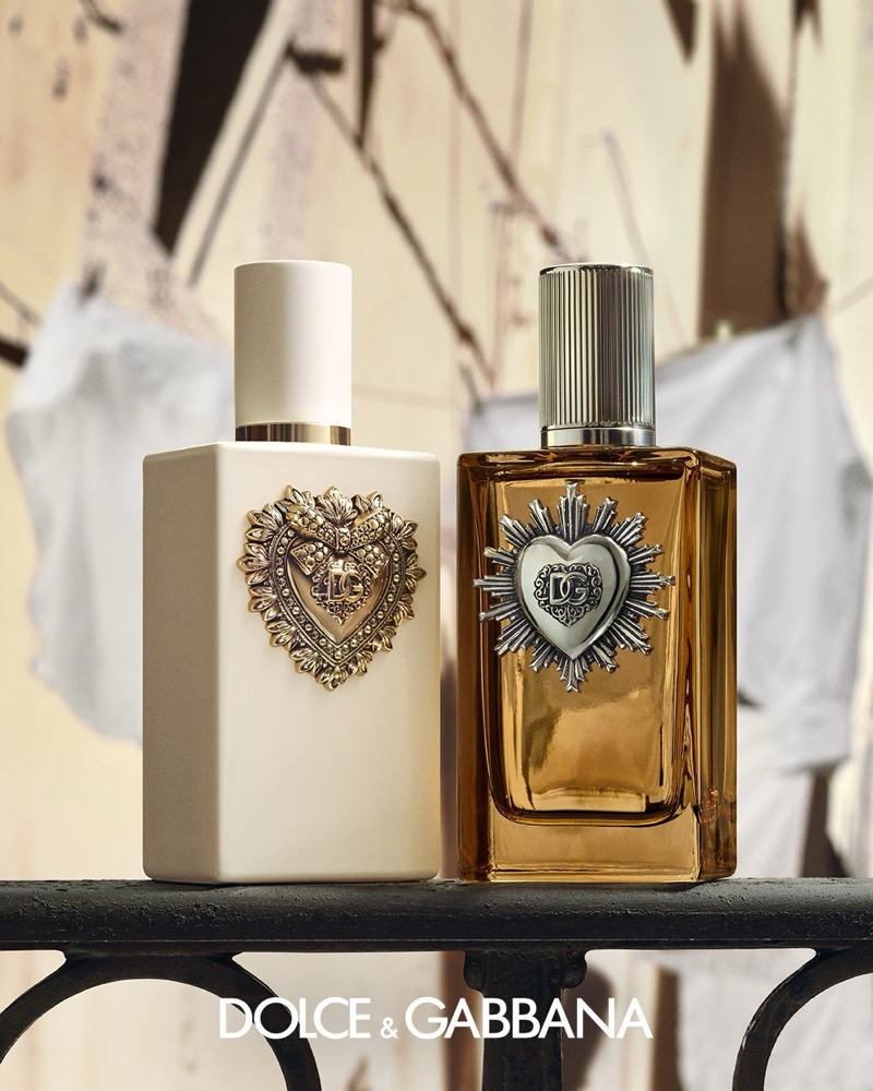 Two Dolce & Gabbana Devotion fragrance bottles on a wrought-iron railing, one with a gold heart emblem and one in amber glass with a silver heart and sunburst