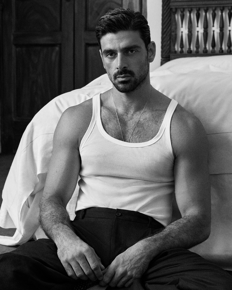 Black-and-white portrait of Michele Morrone in a ribbed tank and a thin chain necklace, seated on the floor against a bed with a carved wooden headboard
