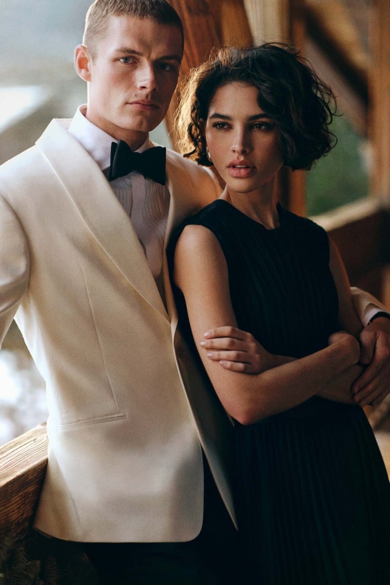 Finley Prentice wears a cream dinner jacket, pleated shirt, and black bow tie beside a woman in a sleeveless dress.