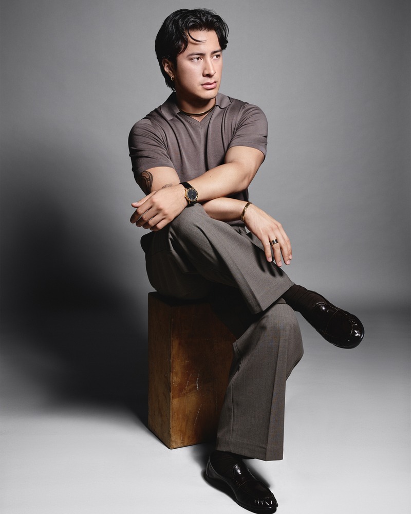Hudson Williams sits on a wooden block in a polo and tailored trousers with Bvlgari jewelry and a watch.