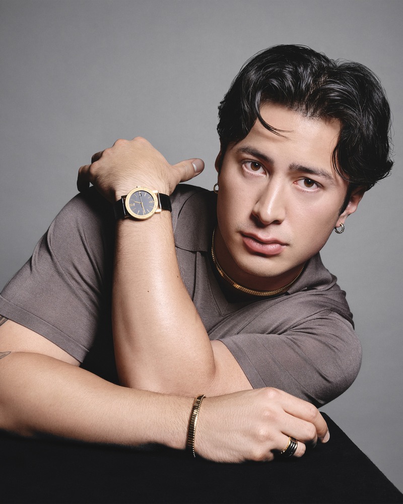 Hudson Williams holds his wrist beside his face, showing a Bvlgari dark-dial watch, necklace, earrings, bracelet, and rings.