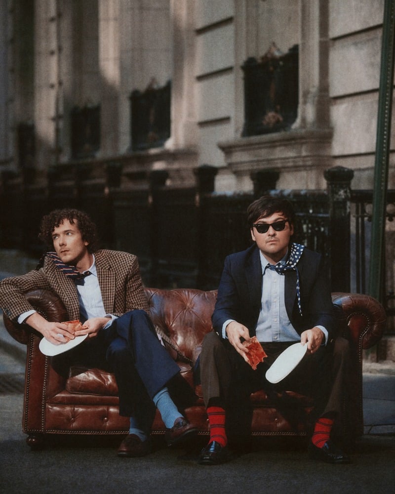 Martin Herlihy and John Higgins hold paper plates while sitting on a leather sofa outdoors.