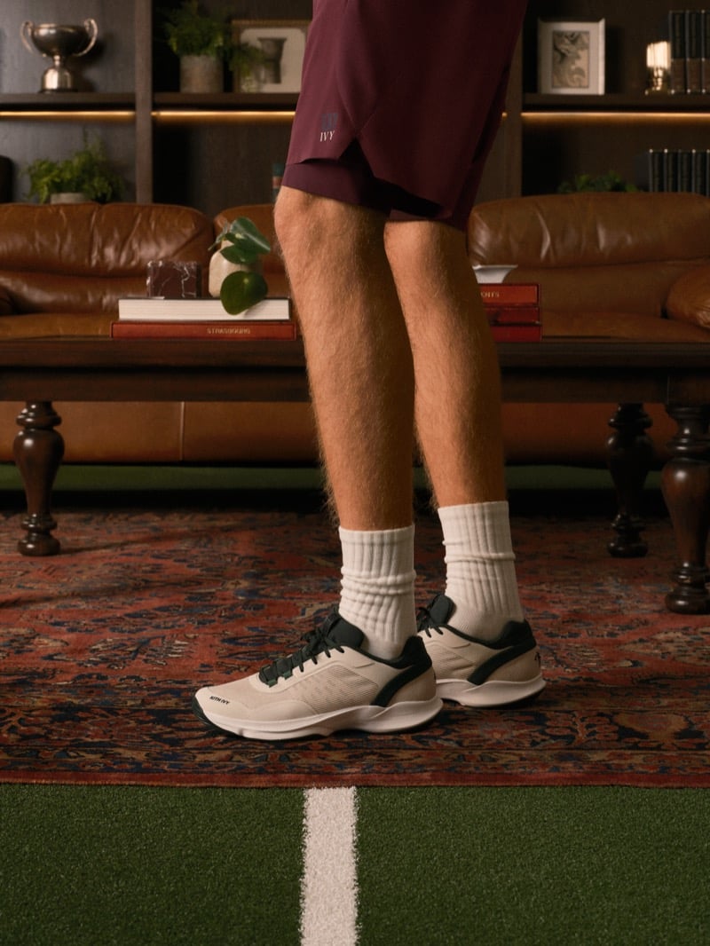 Cropped view of burgundy shorts with an Ivy logo, white crew socks, and light court sneakers with dark trim, standing on a rug beside an indoor turf line
