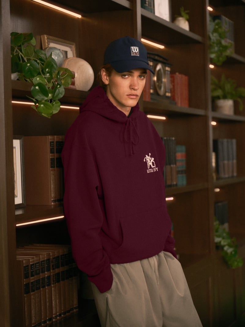 Warren Axelman leaning against library shelves in a baseball cap and a burgundy hoodie with an embroidered Kith Ivy logo, one hand in his pocket
