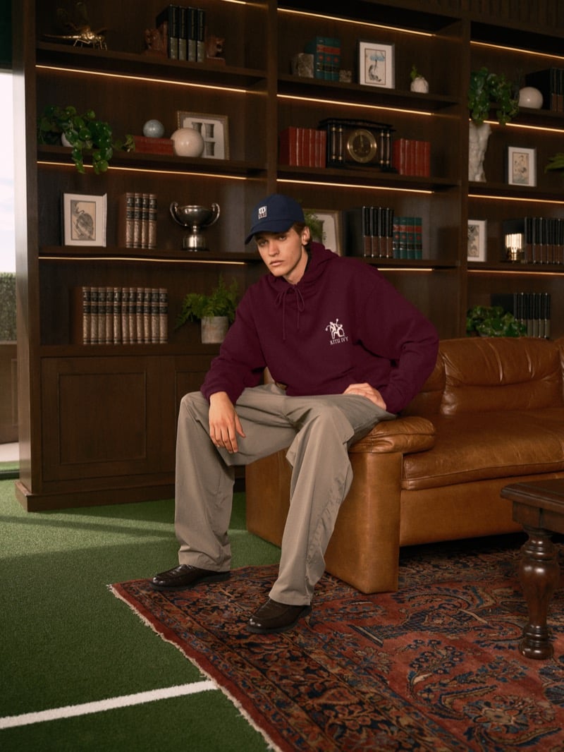 Warren Axelman seated on a leather sofa in a baseball cap, a burgundy hoodie with a Kith Ivy logo, wide beige trousers, and brown loafers on a patterned rug