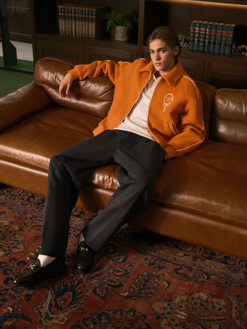 Warren Axelman reclining on a brown leather sofa in an orange varsity jacket over a ribbed polo, wide-leg trousers, and black loafers with gold hardware, a patterned rug below