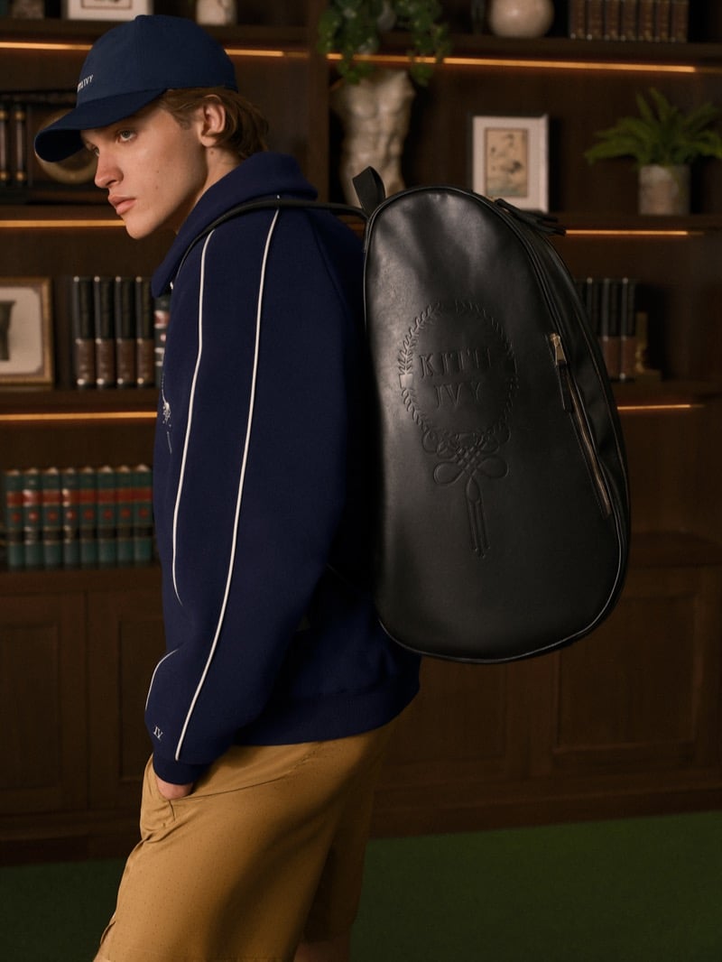 Warren Axelman in profile in a baseball cap and a piped zip jacket, a black leather padel backpack embossed with a Kith Ivy crest on his back, tan shorts below