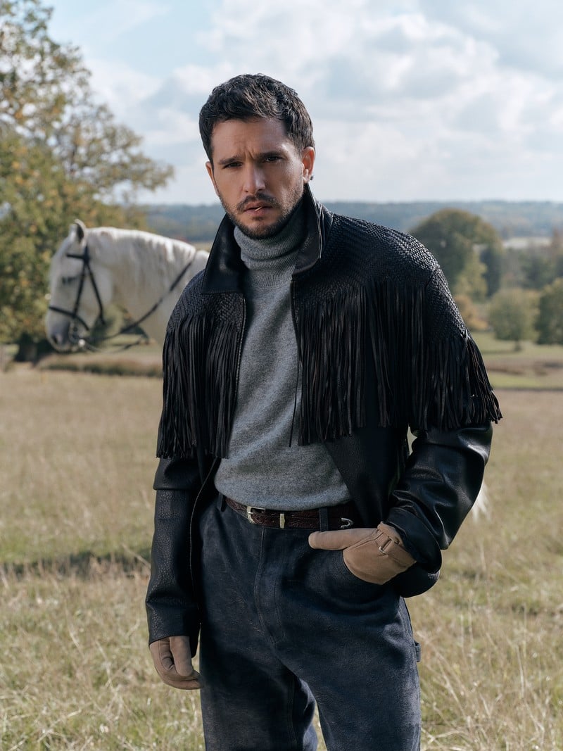 Kit Harington wears a black fringed jacket, gray turtleneck, and dark denim in a field.