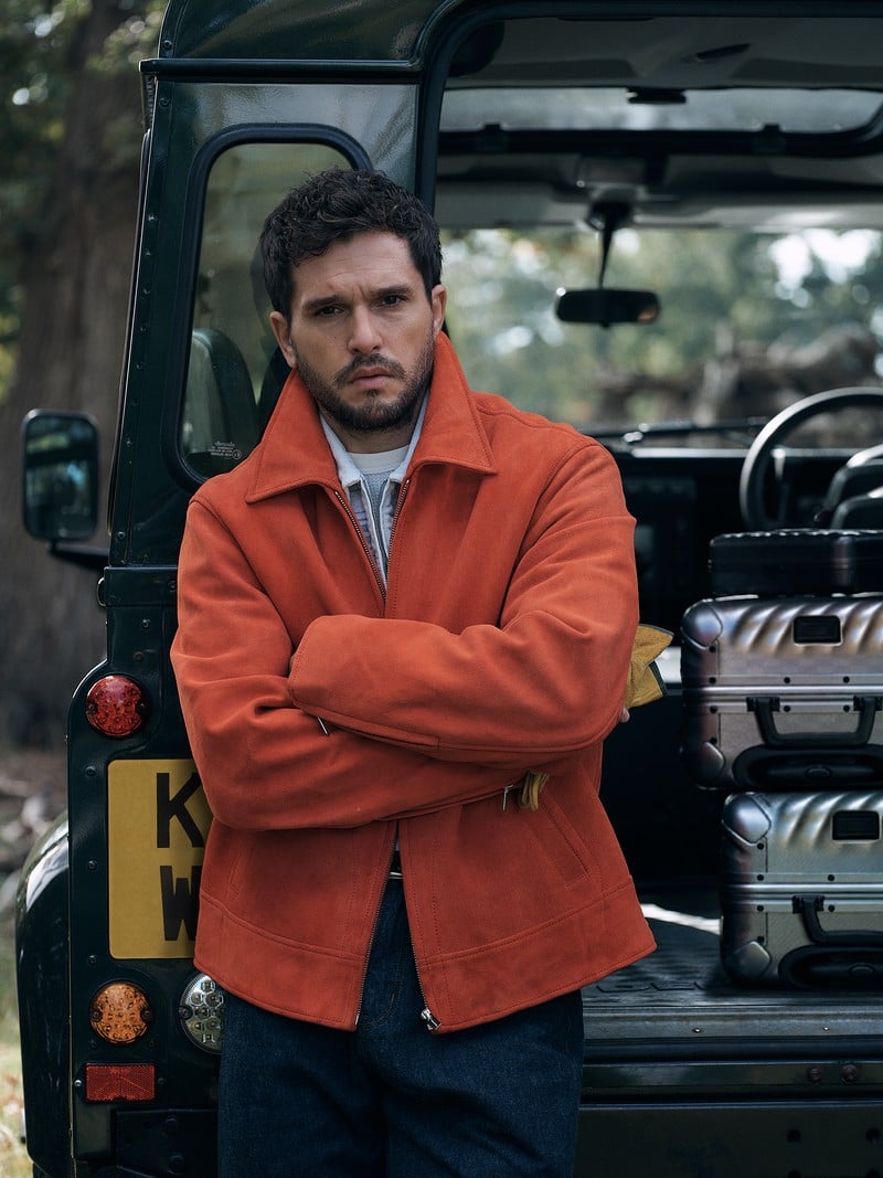 Kit Harington stands beside an off-road vehicle wearing an orange jacket over a denim shirt.