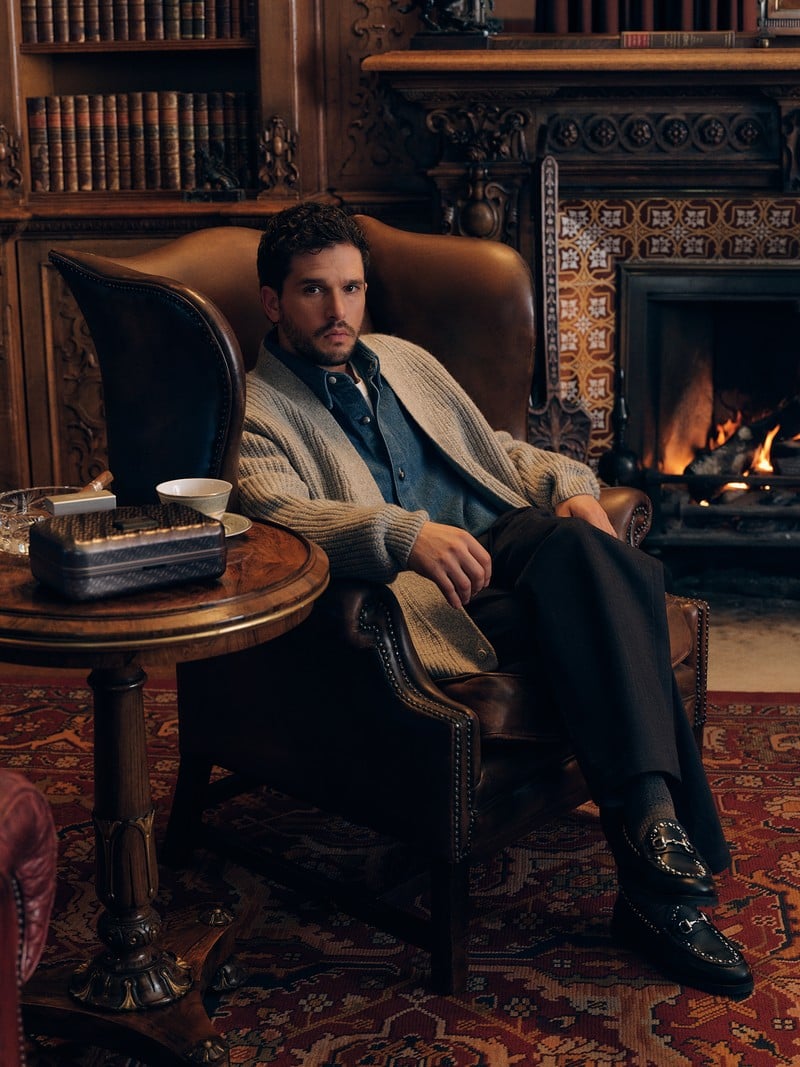 Kit Harington wears a cardigan, collared shirt, dark trousers, and loafers beside a library fireplace.