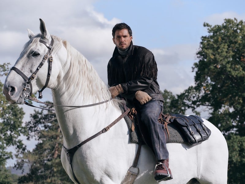 Kit Harington rides a white horse wearing a black fringed jacket, gray turtleneck, and denim.