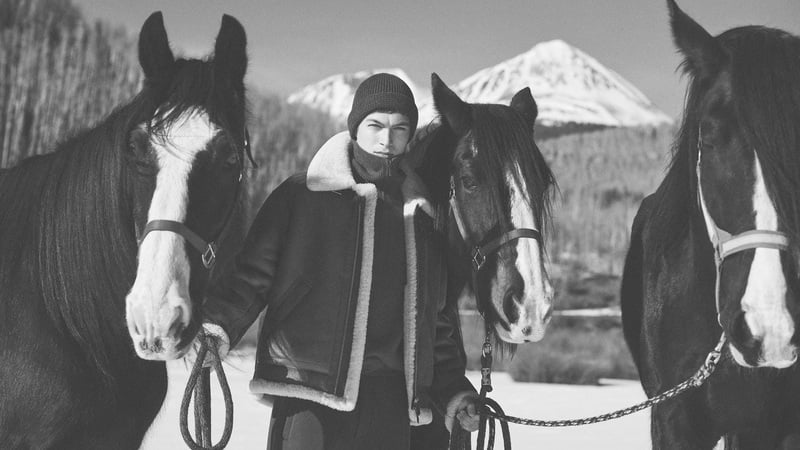 Just Verhoeff stands among horses in a shearling-trimmed jacket and knit cap against snow-covered mountains.