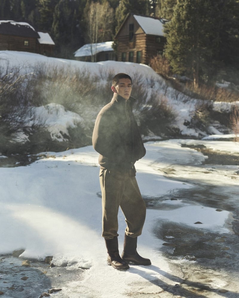 Just Verhoeff stands in snow near wooden chalets wearing a high-collared sweater, trousers, and boots.