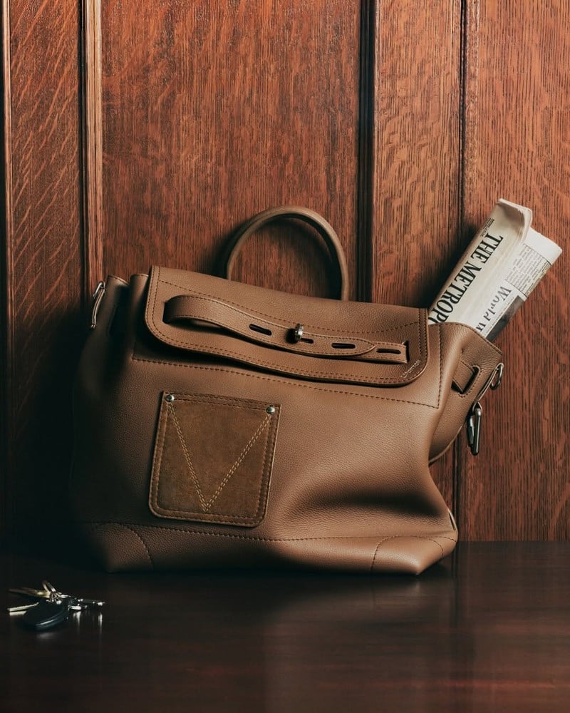 Louis Vuitton leather bag with a buckled strap, V-stitched front patch, and folded newspaper in the Trunk Edition campaign.