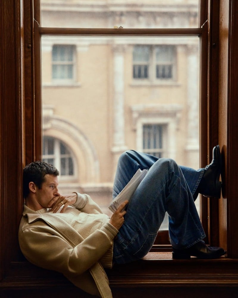 Callum Turner reads in a window seat wearing a jacket, knitwear, jeans, and boots in the Louis Vuitton Trunk Edition campaign.
