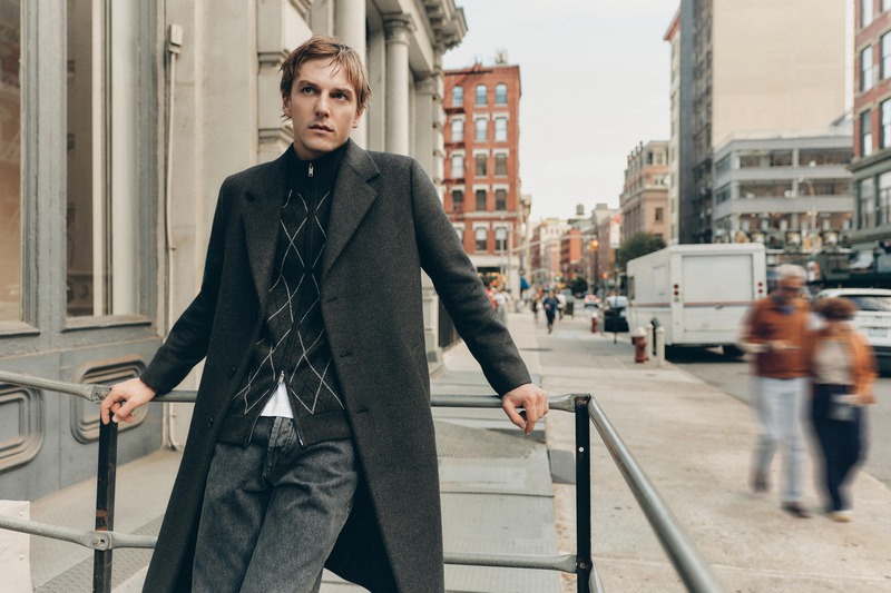 Quentin Demeester leans against a railing in a charcoal coat over a diamond-patterned zip knit and faded jeans for Massimo Dutti's fall 2026 New York edit.