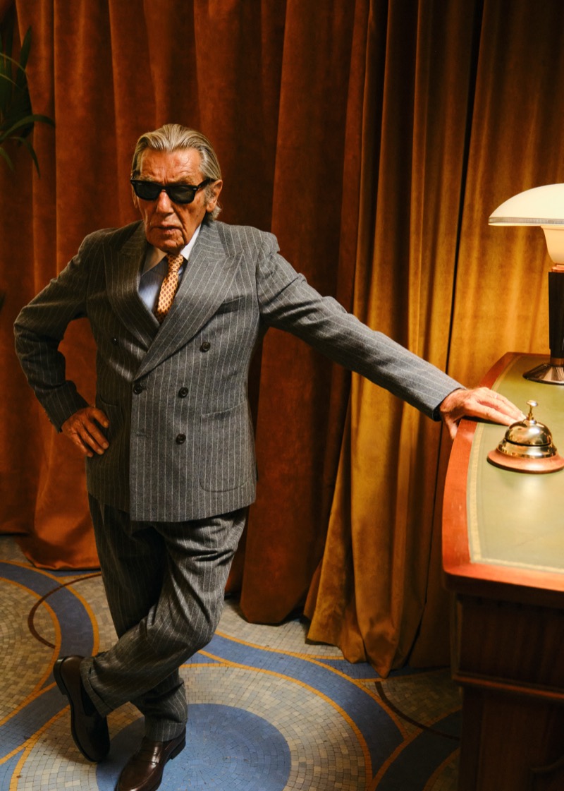 Andrés García-Carro, known as The Spanish King, in sunglasses and a gray double-breasted pinstripe suit leaning on a reception desk beside a service bell