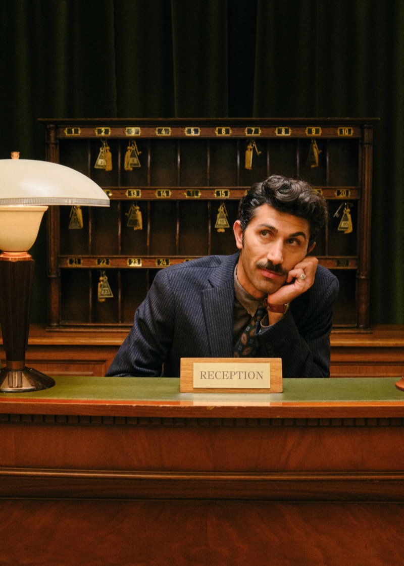 Ata De Çin resting his chin on his hand at a reception desk in a pinstripe jacket and patterned tie, with room keys on the wall behind him
