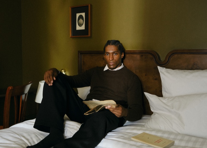 Joe reclining on a hotel bed in a brown sweater with a collared shirt underneath, an open book in his hand