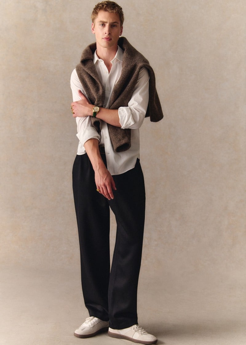 Max Turland wears white Halrin sneakers with dark trousers and a sweater over his shoulders.
