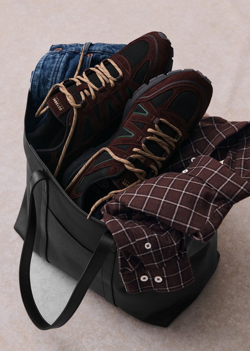 Brown and black OE10 sneakers sit beside folded clothing and a bag.