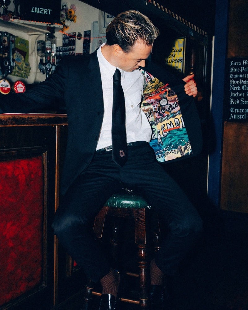 Ned Green of Legss opens his dark jacket to reveal a colorful printed lining while seated on a pub stool.