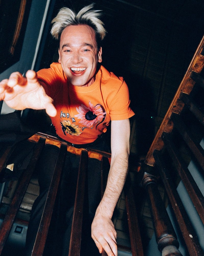 Ned Green of Legss reaches toward the camera wearing an orange sunflower graphic T-shirt.