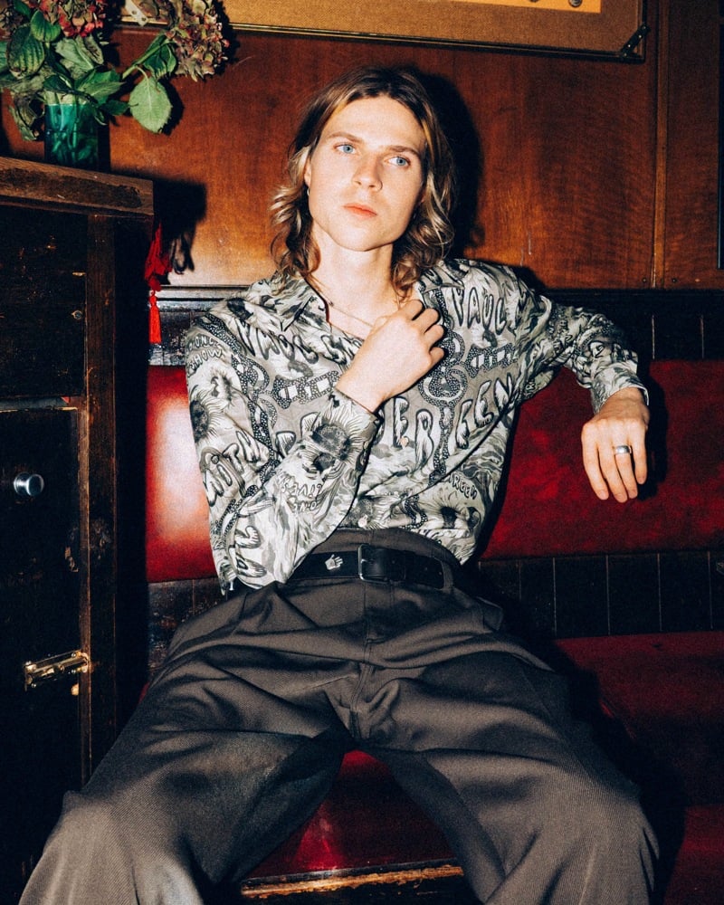 A member of Legss sits on a red pub banquette wearing a patterned shirt and dark trousers.
