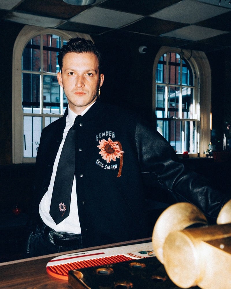 Jake Martin of Legss wears a black sunflower varsity jacket over a white shirt and dark tie.