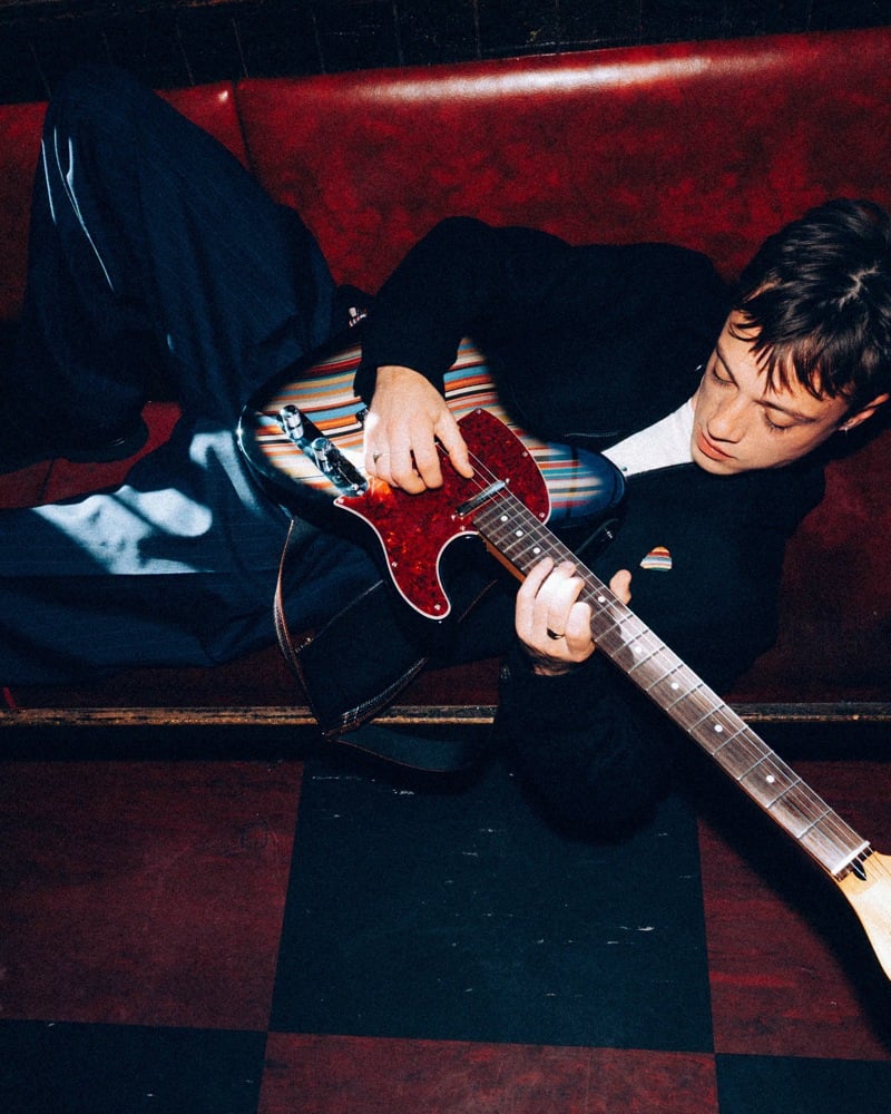 Max Oliver of Legss reclines on a red banquette playing a striped Fender Telecaster.