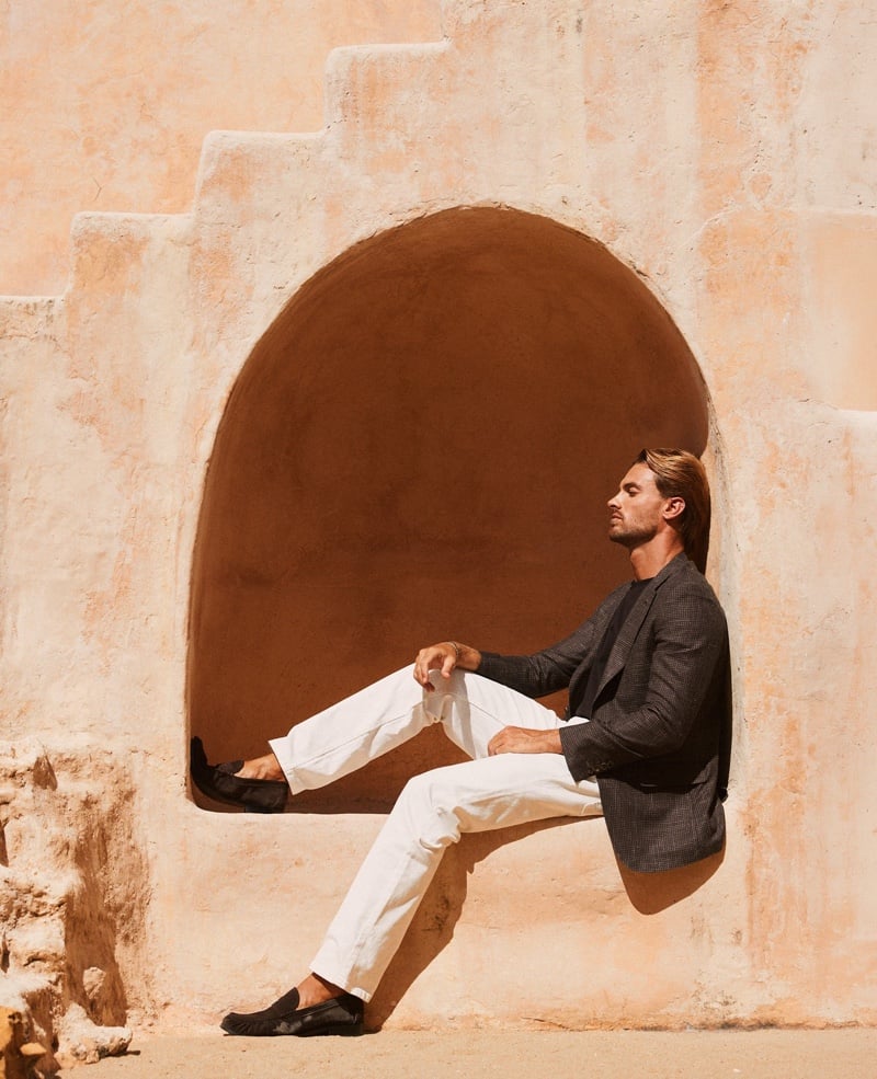 Gonçalo Campos sits in an arched alcove wearing a dark blazer, T-shirt and white trousers with black loafers.