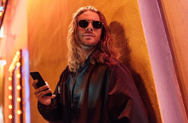 Man with long hair and sunglasses in a black leather jacket holding a smartphone against an orange wall.