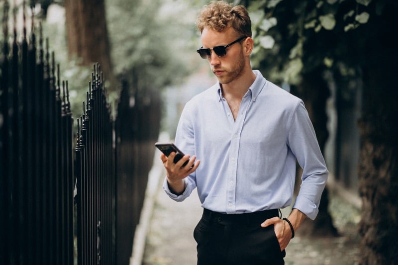Man wearing sunglasses, a pale blue shirt and dark trousers looking at his smartphone outdoors.