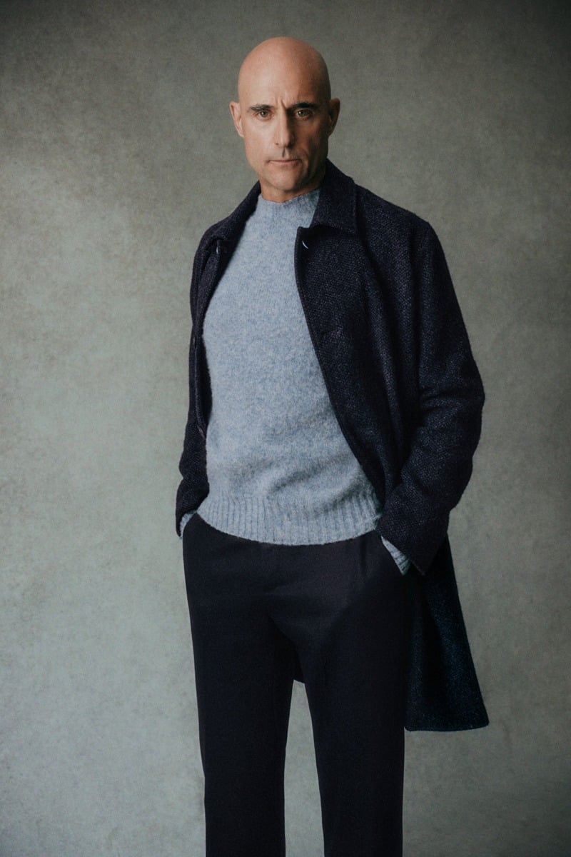 Mark Strong wears a dark coat over a pale blue sweater and dark trousers.