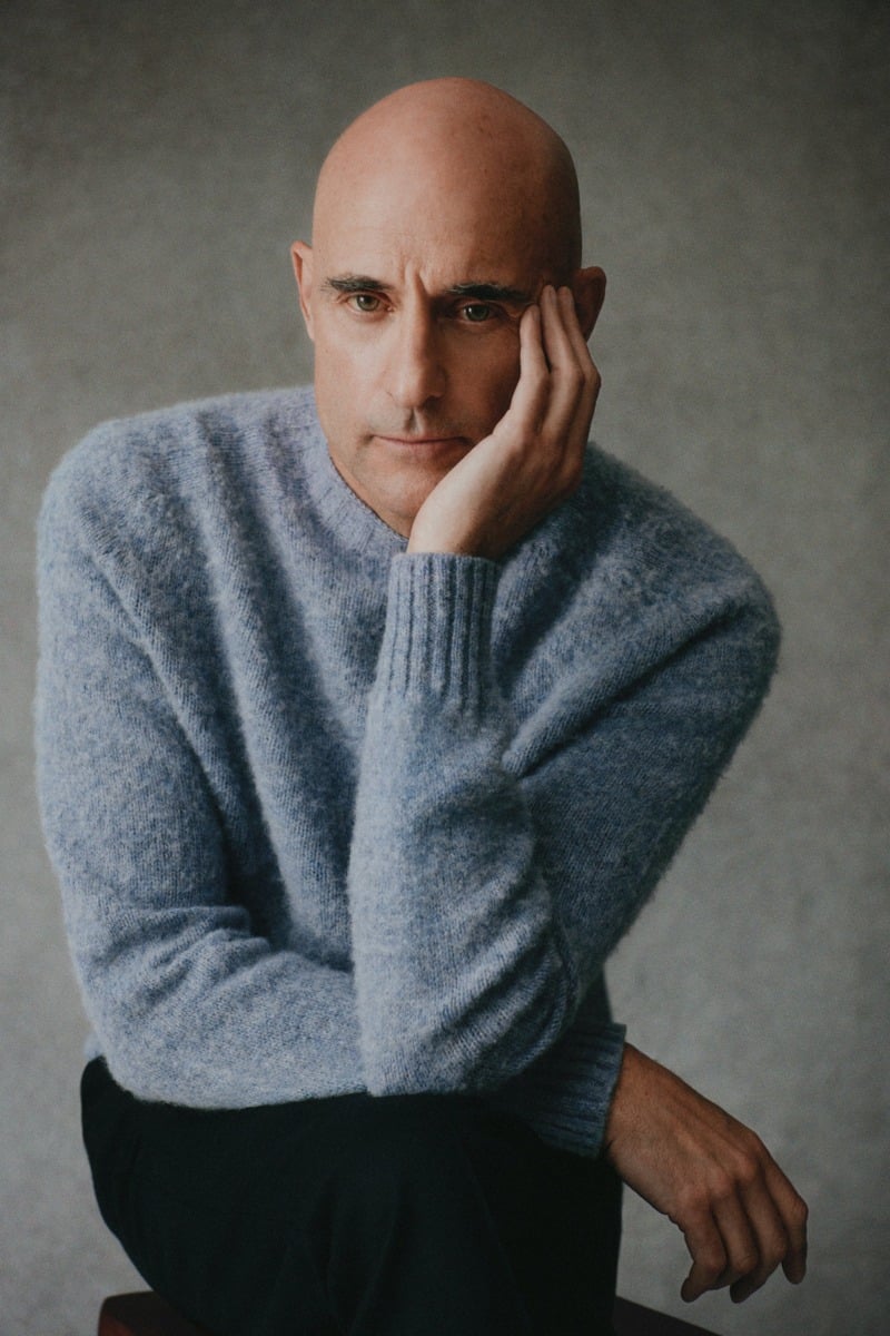 Mark Strong wears a pale blue sweater while resting his cheek in his hand.